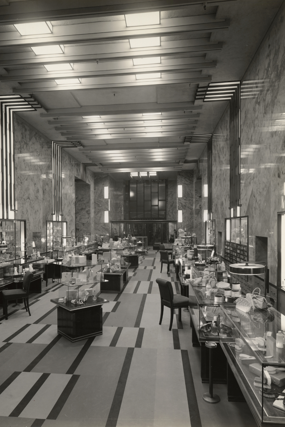Interior of Bullock's Wilshire building, circa 1935. The first-floor cosmetics and perfume department features a high Art Deco ceiling with geometric light fixtures, matching carpet patterns, and marble walls. Display cases line the walls and center of the room showcasing luxury cosmetics, perfumes, and accessories, with chairs placed for customers.