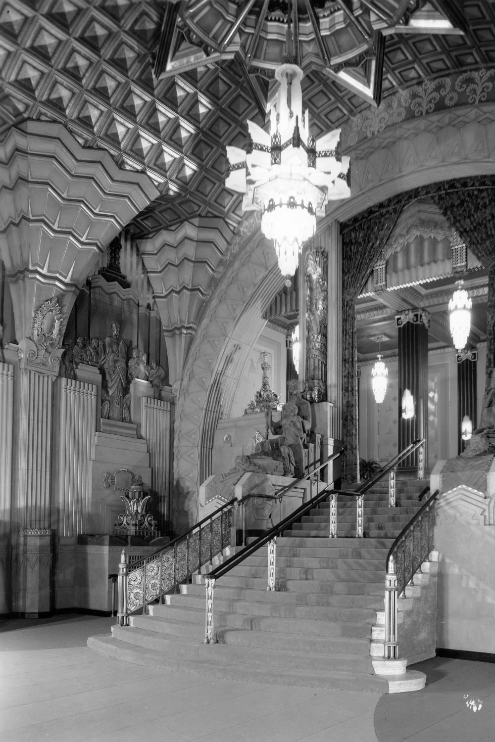 Mezzanine stairway descending into the lobby of the Pantages Theatre, Hollywood, California, photographed in 1929, showcasing ornate Art Deco details with patterned walls, column-supported vaulted ceilings, chandeliers, and statuary.